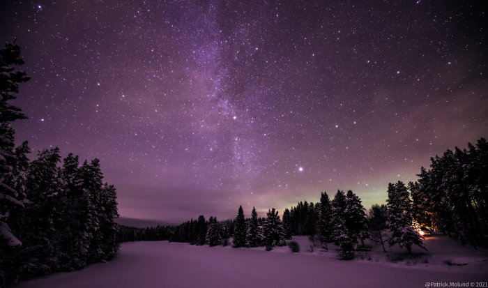 Stjärnklar himmel över snötäckt landskap och träd, med diffust ljus i bakgrunden.