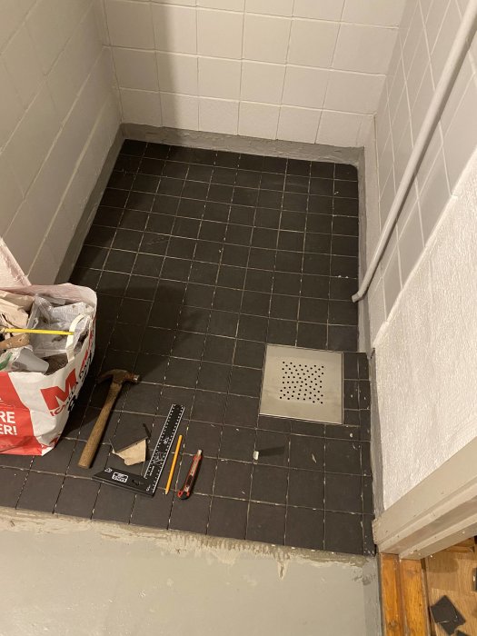 Renoverat badrumsgolv med svart klinkers, ny sil över brunn och byggmaterial samt verktyg på golvet.