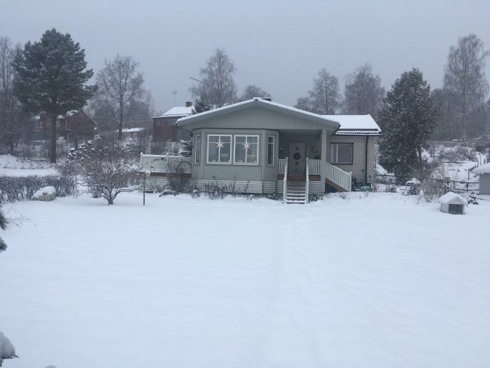 Enplanshus med utbyggnad och altan täckt av snö, omgivet av vinterklädd trädgård.