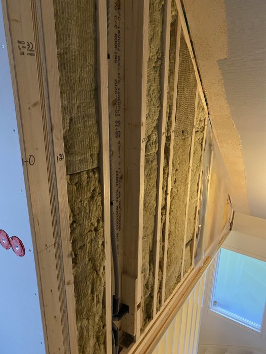 Nyinstallerad isolering mellan träreglar i en vägg med synliga elinstallationer och en takfönster.