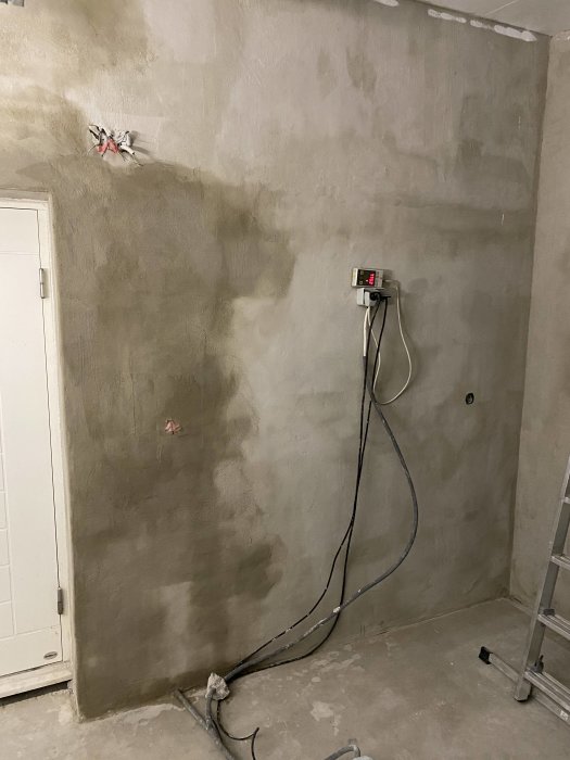 Delvis putsad garagevägg med synlig elinstallation, kablar och en dörr. Skuggor syns på väggytan.