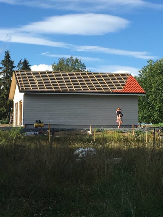 Byggnad under konstruktion med liggande panel och takstomme, en del takpannor lagda, och en kap- och geringssåg framför.