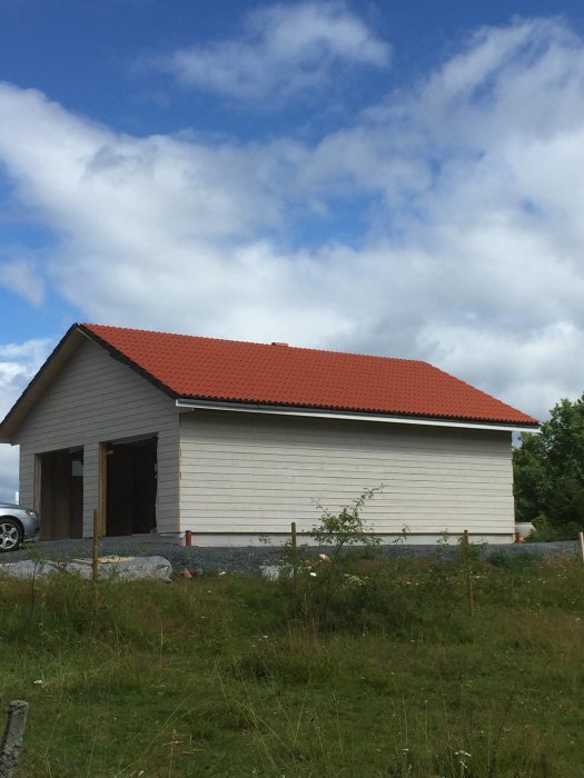 Nybyggt hus med rött tegeltak, beige panel och öppen garageport under en blå himmel med moln.