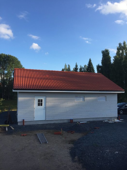 Vitputsad byggnad med rött tegeltak och liggande panel under en klarblå himmel.