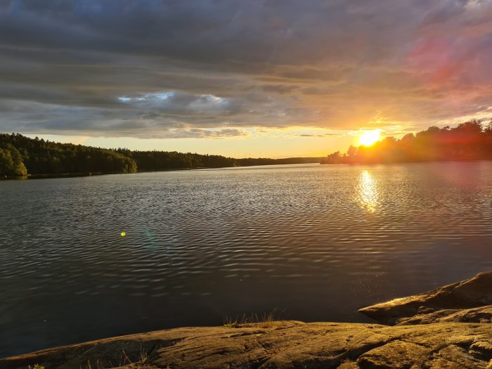 Solnedgång vid en sjö med ljusstrimma på vattenytan, sedd från en klippa.