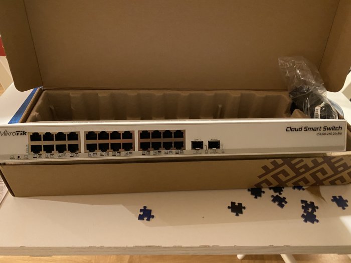 Ny MikroTik Cloud Smart Switch i öppen kartong på ett bord, bredvid pusselbitar.