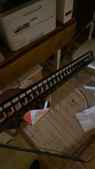 Patchpanel hållen i handen med synliga numrerade portar, ovanför ett rörigt trägolv fullt med kablar och en låda.