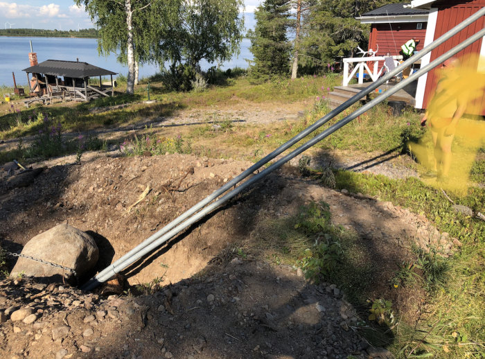 Grävplats med ett stort hål och en stor sten, en metallstång fastkedjad till stenen för bortflyttning, i skärgårdsmiljö.