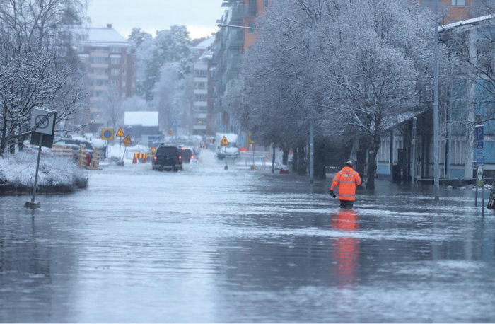 Översvämmad gata med person i arbetskläder, vattensamlingen och vinterträd i Huddinge.