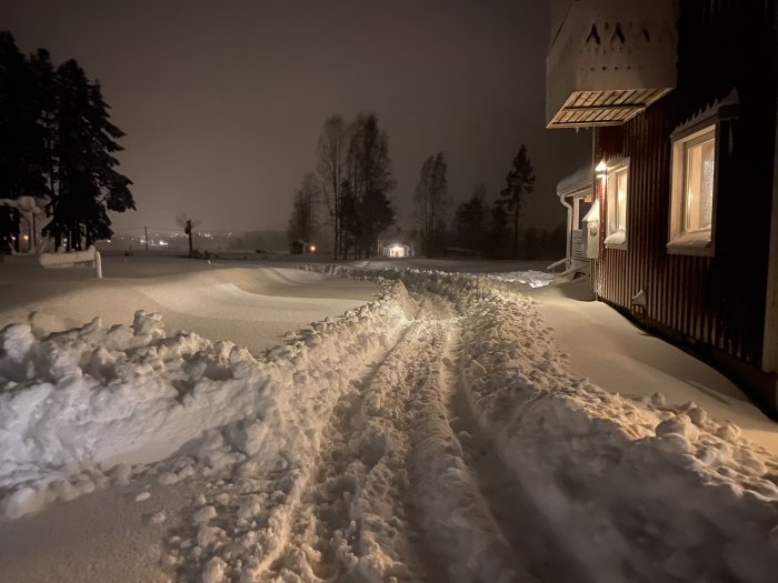 Snötäckt uppfart på natten med uppkörda hjulspår som leder till ett hus med tänd utomhusbelysning.