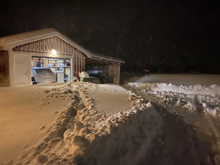 Garage med öppen dörr och synligt innehåll, med fotspår i snön framför under kvällstid.