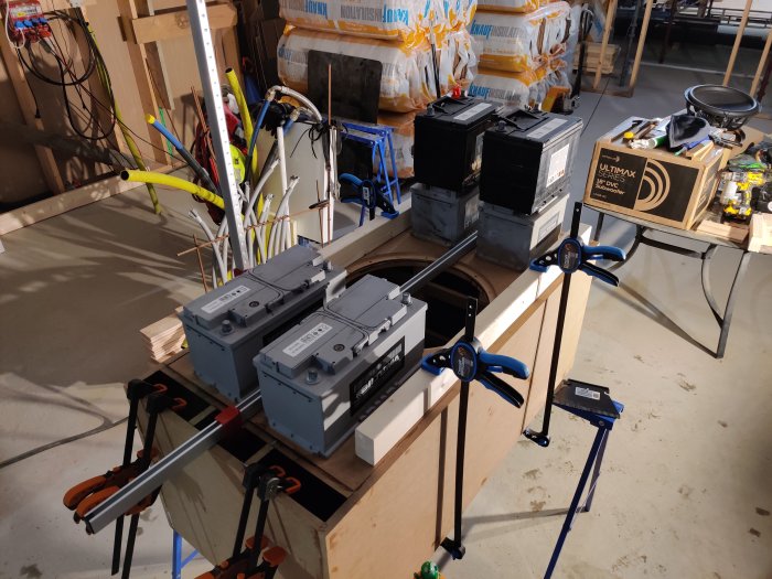 Hobbyverkstad med byggmaterial, högtalarelement på kartong och flera båtbatterier på ett arbetsbord.