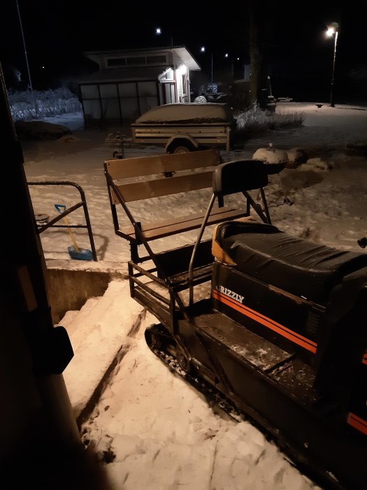 Snöskoter med ett nytillverkat träbaksäte på pakethållaren för barnens säkerhet, i ett snötäckt landskap på kvällen.