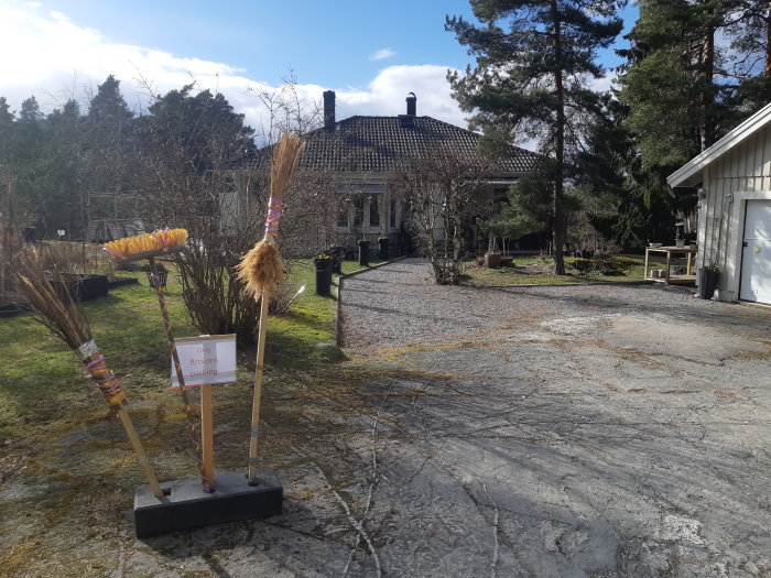 Flertal kvastskaft står på en uppfart med skylten "Only Brooms parking" framför en villa och garage.