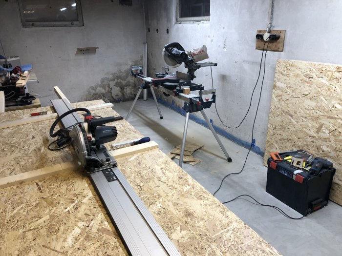 Delvis renoverat källarutrymme med verktyg och kapsåg på ett arbetsbänkssystem, osb-skivor på väggen och rör på väggen.