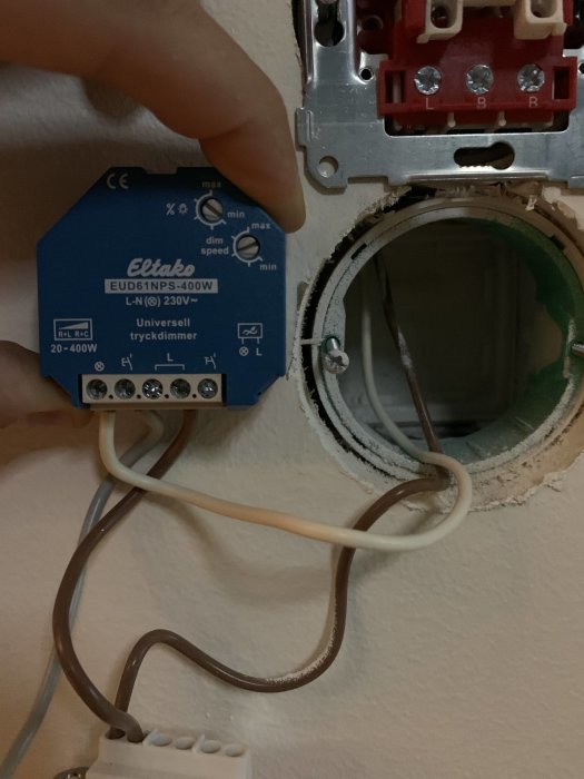 En hand som pekar på en blå Eltako universell tryckdimmer installerad bredvid en öppen eldosa i en vägg.