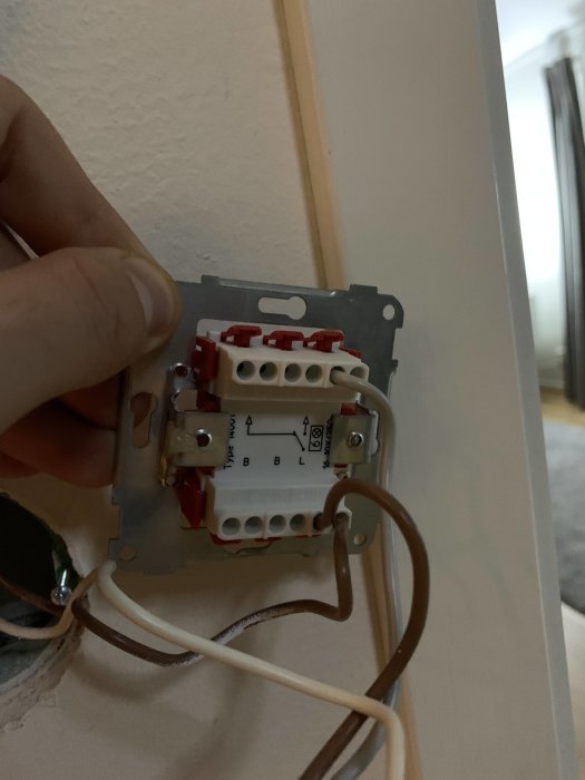 Hand som håller en öppen väggdimmer med synliga ledningar och terminaler mot en vit vägg.