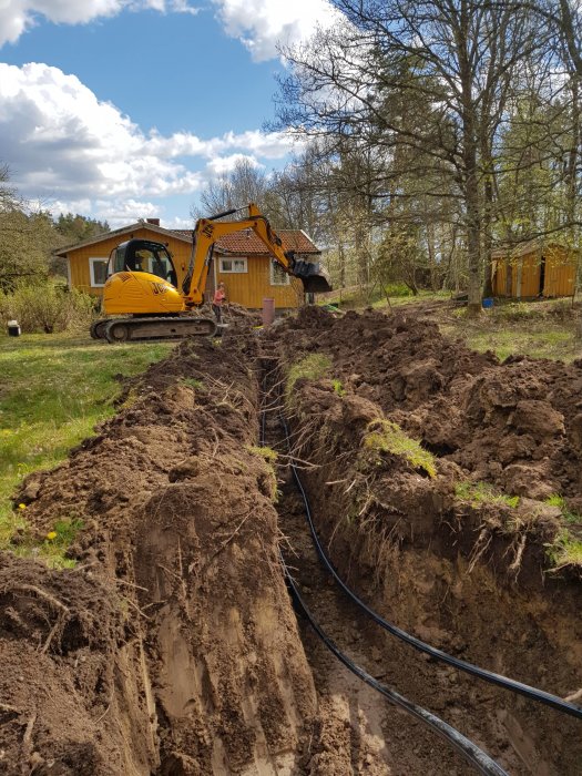 Grävmaskin gräver en djup schakt för förberedelse av bergvärmeinstallation på en tomt med grönska.