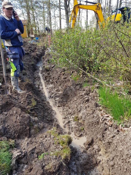 Man i arbetskläder står bredvid grävd schakt för bergvärmeinstallation med grävmaskin i bakgrunden.