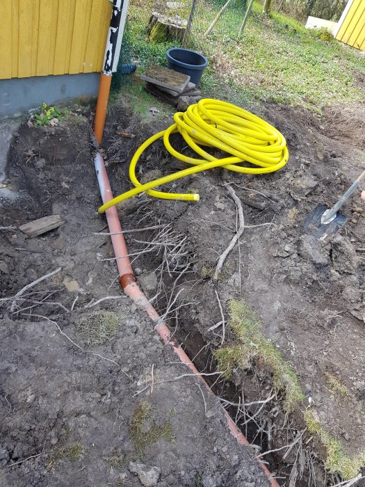 Grävd trädgård med synliga rörledningar och en gul slinga av bergvärmeslang på marken.