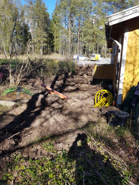 Uppgrävd trädgård med slangar för bergvärme nära en byggnad, träd och parkerade bilar i bakgrunden.