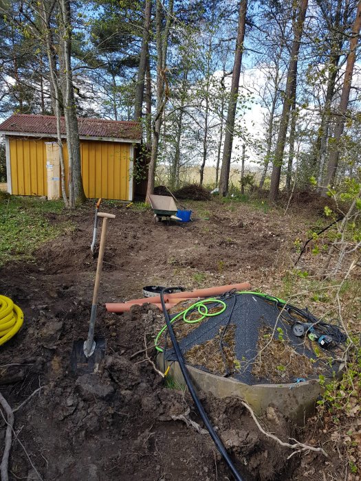 Uppgrävd trädgård med verktyg framför ett litet gult skjul, förberedelse för bergvärmeinstallation.