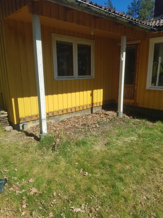 Gul trähusfasad med borttagen veranda, förberedd för framtida utbyggnad, synlig gräsfundament.