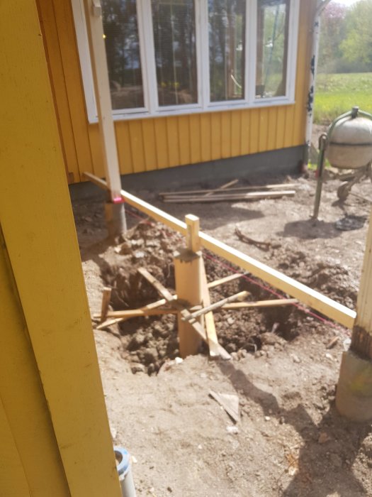 Grävplats för utbyggnad nära hus med synliga fundament och demonterad veranda i bakgrunden.