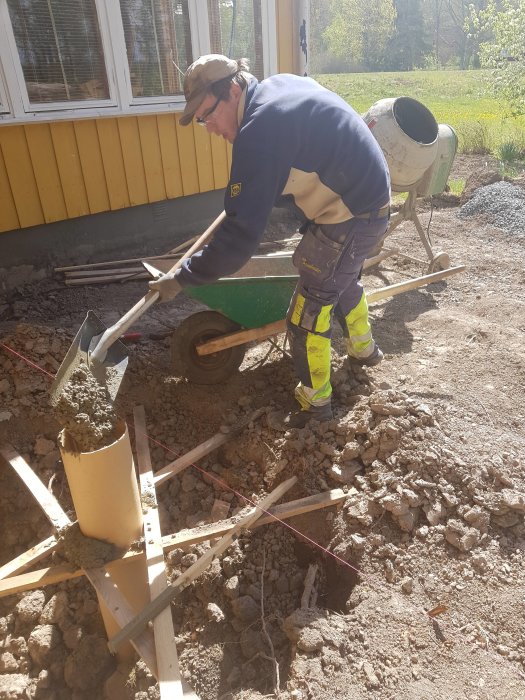 Arbetare häller betong för grunden till en utbyggnad nära hus med gul fasad.