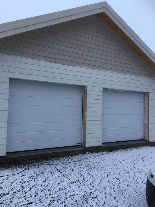Nybyggda garaget med vita dubbeldörrar och obehandlat trä runtom, med ett lätt snötäcke framför.