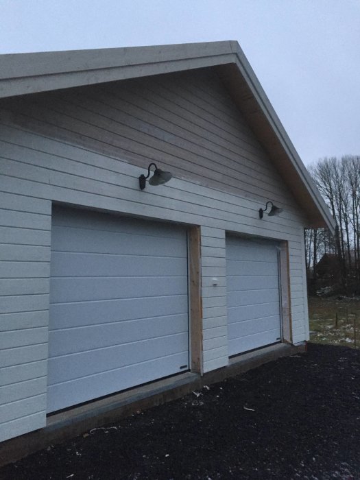 Nybyggt garage med två grå portar, vit plåtklädsel runtom och två utomhuslampor monterade ovanför portarna.