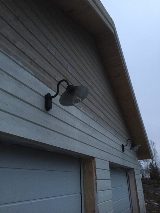 Uppvinklade vägglampor monterade på en träbeklädd yttervägg ovanför ett garage i skymningen.