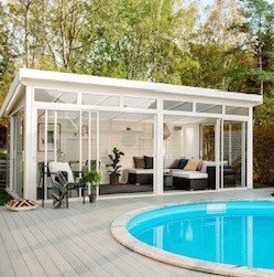 Vit poolhus i Friggebodsstorlek nära en rund pool omgiven av trätrall.