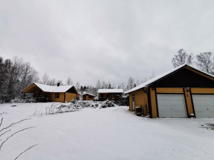 Snöklädd tomt med gula hus och en dubbelgarage i förgrunden och nyligen avverkade trädområden i bakgrunden.