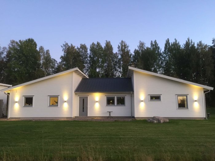 Ett nybyggt 1-plans Varbergshus med vit fasad och mörkt tak i kvällsljus, upplyst av utomhuslampor.