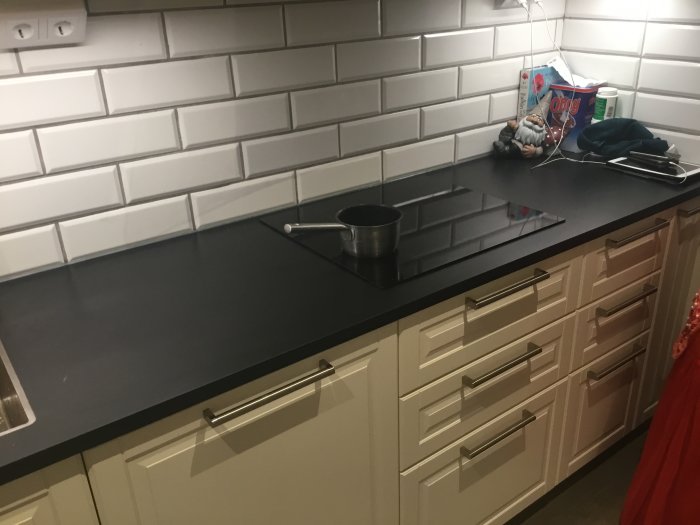Kök med svart bänkskiva, beige skåpluckor och vit tegelliknande kakelvägg.