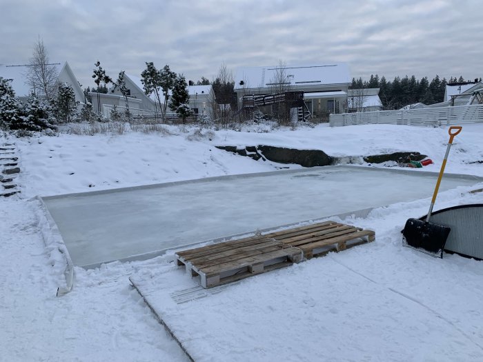 Hemmagjord isbana med upphöjda kanter av trä och plast i en snöig trädgård, skottkärra och snöskyffel syns.