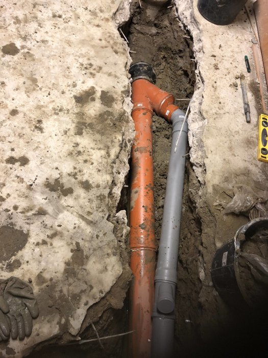 Rörinstallation i grävd ränna med orange och grå dagvattenledningar och verktyg på en betonggolv.