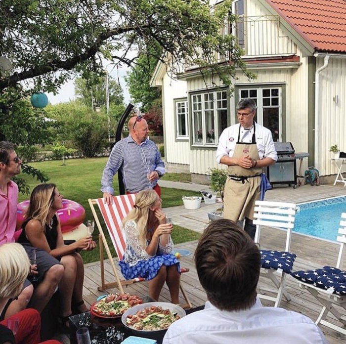 Grupp människor njuter av en grillkväll vid poolen utanför ett hus, i en sommarträdgård med mat och dryck.