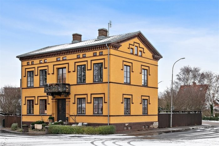 Tvåvåningshus i gult tegel med snötäckt tak och svart smidesjärnsbalkong, omgivet av ett snöstrött landskap.