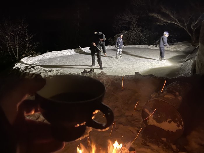 En person håller i en kopp nära en eld, med en bakgrund av människor som sopar snö på en upplyst isbana om kvällen.