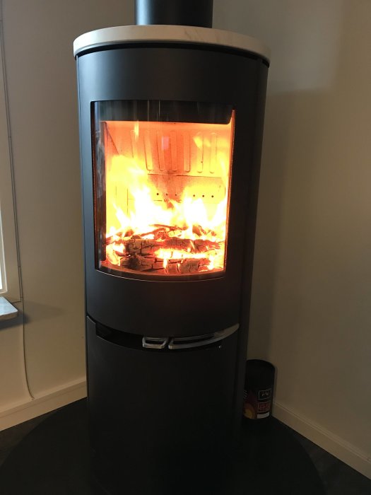 Modern kamin av modellen Aduro H2 med eld och brinnande ved inuti, belyst från sidan.