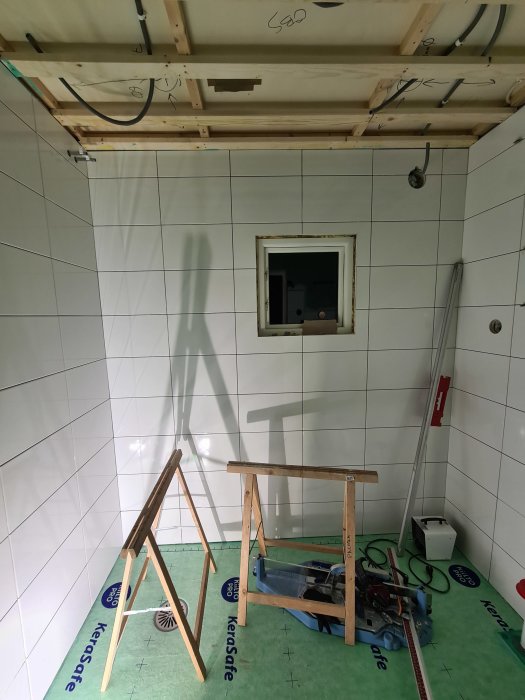 Badrum under renovering med kaklade väggar, osatt tak, byggsågar och arbetspall.