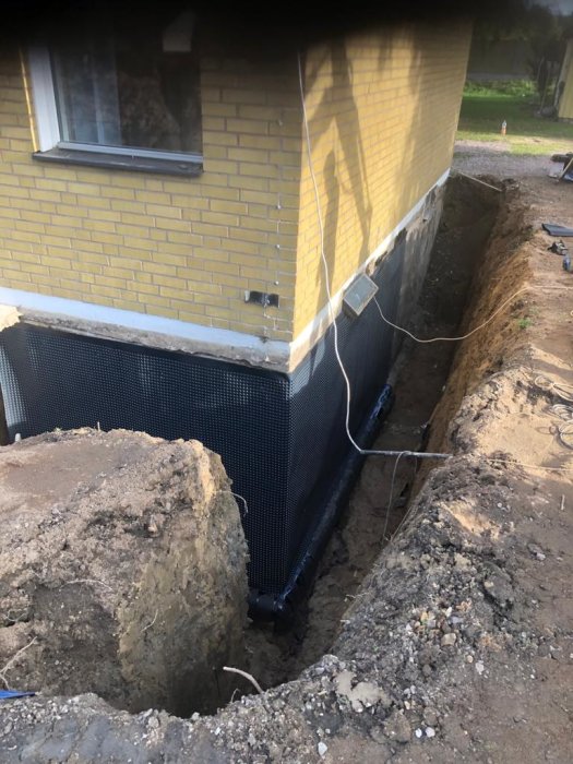 Grävd dräneringsdike vid husgrund med installerad dräneringsrör och skyddande geotextil.