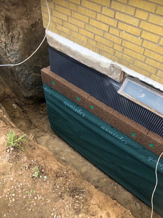 Dräneringsarbete vid husgrund med geotextil och dräneringsplatta vid gult tegelhus.