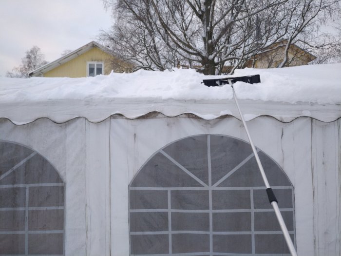 Takskottning med snöskovel på långt skaft framför ett snötäckt tält och ett gult hus i bakgrunden.