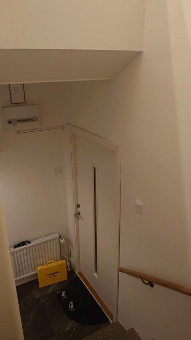 Ett trapphus med vitmålade väggar, en ytterdörr, en radiator och en installerad luftvärmepump ovanför dörren.