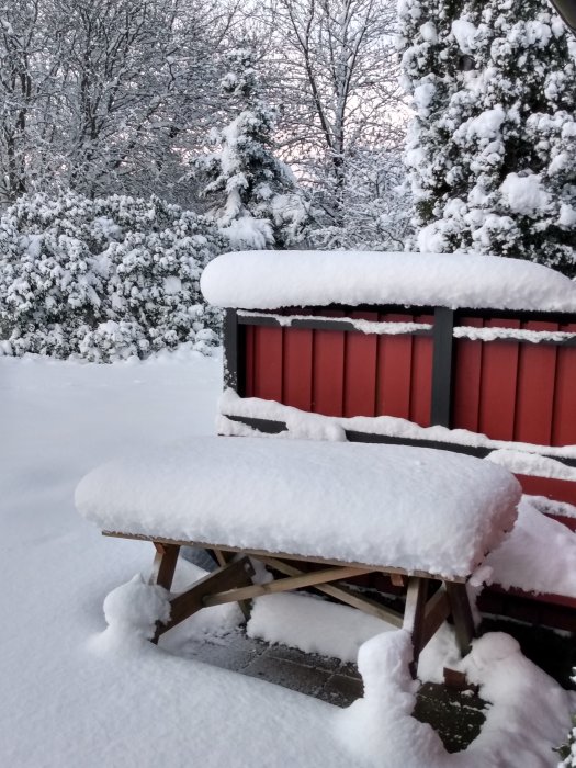 Snötäckt trädgård med tjockt lager vit snö på bord och bänk, träd i bakgrunden.