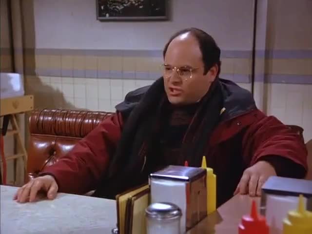 Man i röd jacka och glasögon sitter vid ett dinerbord ser irriterad ut.
