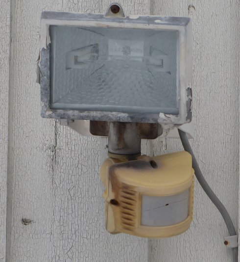 Gammal strålkastare med sprucken och bränd rörelsedetektor monterad på vit vägg.